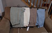 Four pairs of pants laid on a couch horizontally on hangers, showing the elastic waistbands and colors - light green, white, taupe, light blue.