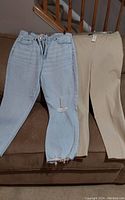 Two pairs of pants laid out on a couch, one light blue distressed denim with rips and frayed hems and one beige pair with tags attached.