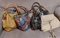 Overview photo showing the five purses arranged on a beige couch: tan leather handbag with flap and tassel, colorful floral-paisley fabric tote, braided handle dark leather purse with studs, camel leather purse, and tan leather purse.