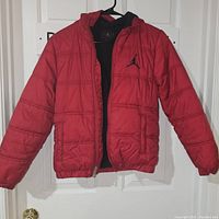 Front view of red Jordan kids' puffer jacket hanging on black hanger showing the jacket's quilted design and Jordan logo on chest.