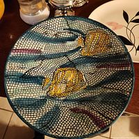 Round serving plate with mosaic-style pattern of netted blue, gray background and two yellow seashells, placed on a wooden surface with a silver decorative item behind.