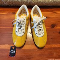 Top front view showing yellow leather and suede sneakers with white laces and flower charm on one lace.