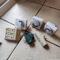 Front view of three mechanical timers, beige 4-outlet splitter, and two plug adapters on tiled floor