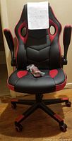 Front view of gaming chair with red and black upholstery and padded armrests raised. Instruction sheet attached to the chair back and spare caster wheels on the seat.