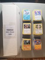 Top-down view of multiple stacks of assorted Pokémon trading cards next to a labeled white storage box.