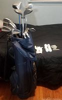 Full view of golf bag standing upright with clubs inside and golf balls, tees, and gloves laid out on bed next to it.