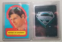 Photo showing front and back of two Superman The Movie insert stickers from 1978 Topps Series 1. The front sticker is a portrait of Superman with a bright blue background and red border. The reverse shows a silver Superman logo on black.