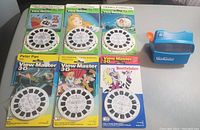 Photo showing six 3D reels in original opened packaging laid out alongside the blue TYCO Model L View-Master viewer.