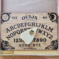 Top view of the Ouija board showing letters, numbers, yes/no, and decorative artwork with the planchette centered on the board.