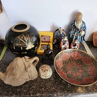 Showing entire lot on granite countertop, including clay pot, black and gold vase, various Asian figurines, Thailand mirror compacts, and brass bowl