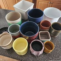 Overview of 14 ceramic and pottery plant pots in various colors, finishes, and shapes arranged on kitchen counter.