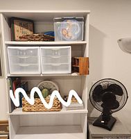 Shelf showing black table fan next to shelves with plastic drawers, baskets, and wooden box