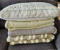 Stack of three folded 100% cotton queen size blankets with a white Oui organic cotton pillow on top. Pillow text shows "STANDARD DOWN ALTERNATIVE PILLOW, 100% ORGANIC COTTON SHELL, 100% RECYCLED POLYESTER FILL".