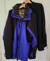 Two men's jackets hanging on hangers, front blue/black Gore-Tex jacket with hood visible, black jacket behind