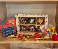 Shelf display with all ornaments including boxed large swirl balls, packaged blue miniature balls, red and gold tinsel picks, and plastic angels.