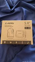 Front of the box showing product details such as frequency 2.4GHz, 5.0 inch TFT LCD screen, and icons for features including night vision, temperature, microphone, and lullabies.