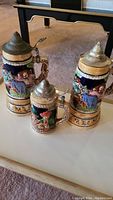 Three ceramic German-style beer steins with colorful relief figures and pewter lids, shown grouped together.