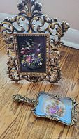 Ornate gold-tone picture frame with floral artwork and hand painted porcelain dish with a pastoral scene, displayed on hardwood floor.