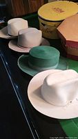 Five assorted hats including three white hats, one beige hat with a pink ribbon, and one green velvet top hat.