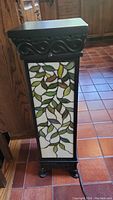 Front view of Tiffany style pedestal lamp showing stained glass leaf design panel with a mix of green, brown, and white glass. Dark colored frame with carved details on top and feet visible.