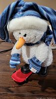 Clear view of the plush snowman with blue and white striped hat, mittens, and orange carrot nose, standing on red skateboard base.