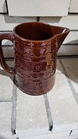 Front and spout view of the brown stoneware pitcher with decorative embossing, placed on a tiled surface.