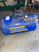 Front side view showing the Power Fist 10 Gallon Portable Air Tank in blue with attached black hose and gauge on concrete floor.