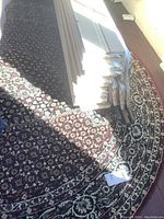 Overall view of the round area rug on floor with a paper tag visible, showing the rug's size and pattern colors including black, red, and beige.