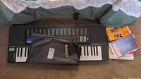 Casio keyboard placed on carpet with black cover partially draped and a pile of music books to the side.