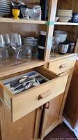Wooden cabinet shelves and open drawer showing ceramic plates, bowls, wine glasses, juice glasses, insulated mugs, and some cutlery.