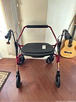 Front view of the maroon folding rollator with a black plastic seat, shown indoors with hand grips and wheels visible.