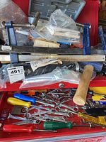 Close-up photo showing large metal F-style clamps, wooden mallet, wrenches, and screwdrivers with colorful plastic handles inside a red toolbox