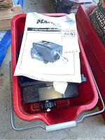 Master Lock 12 Volt electric winch manual inside red plastic pail with winch unit partially visible.