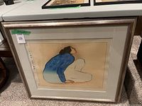 Overall view of framed artwork leaning on table showing silver metallic wood frame, cream mat, dimension tape and MaxSold lot number sticker.