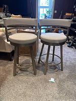 Front view of two matching swivel bar stools with light grey fabric upholstery and metal frames, showing curved backrests and circular footrest rings.