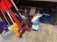 Photo showing brooms, dustpans, vacuum cleaner hose, and blue bucket inside a corner space.
