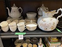 Lot contents showing tea pot, coffee pot, large floral embossed pitcher, cups, saucers, plates, and sugar bowl arranged on a shelf.