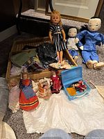 Vintage dolls including Asian dolls, handmade clothes and accessories, and a blue toy case displayed on a carpeted floor.