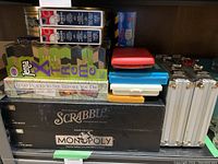 Photo of stacked board games including Scrabble, Monopoly, Jenga Xtreme, and other boxed games, with storage cases on the right side.