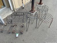 Lot of metal garden accessories arranged on concrete outdoors: several railing baskets, plant stands including round and rectangular, and a small circular ring stand, visible rust and wear.