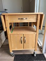 Full view of the wood rolling kitchen cart showing drawer, cabinet with double doors below, drop leaf sides, and caster wheels.