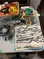 Photo showing folded white cloth tablecloth, matching cloth napkins and place mats with a black branch pattern, plastic and metal cookie cutters, brass candlesticks, clear plastic kitchen food scale, stainless steel popcorn popper, and clear ceramic bowl on a table.