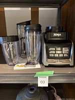 Ninja blender set on shelf showing blender base and three different sized blending cups with lids. Main carafe is missing.