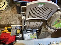 Beige resin patio chair with slatted backrest, red emergency first aid kit pouch, red carrying bag with gloves, and part of bocce set carrying case visible in background.