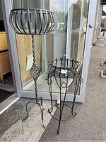 Front view of two metal plant stands with distinct designs and finishes placed outside a building.
