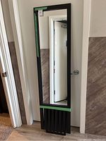 Full view of tall black framed mirror leaning against a wall near a doorway, showing its rectangular shape and lower vertical slat panel design.