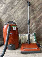 Kenmore orange canister vacuum with hose, metal telescoping wand, power head attachment, and vacuum bags behind it on floor
