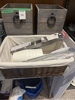 Two square wooden planters with metal ring handles on a black shelf, with a woven basket containing plastic plant stakes and small white ceramic pots inside a white liner.
