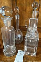 Four clear glass liquor decanters shown together on wood surface with stems and stoppers visible