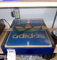 Top view showing Adidas trefoil logo, black hands, yellow markers, and plastic cover taped at sides.